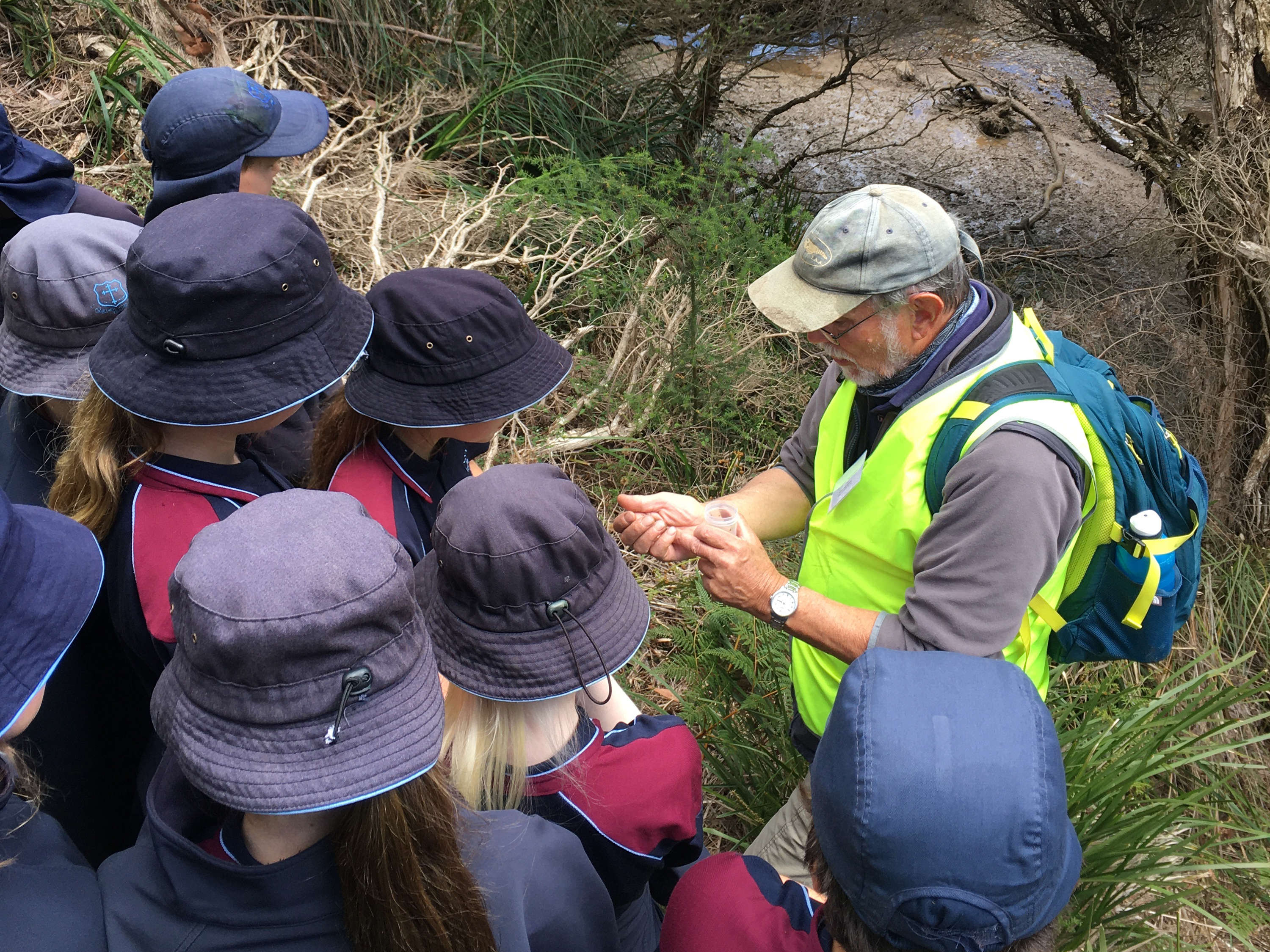 Bookend Trust Academic Director, Alistair Richardson, talking with a group of nine primary school students. They are standing in the bush at the edge of a river. Alistair has specimens in the palm of his hand, which is discussing with the students. The school students are wearing navy blue hats and Alistair is wearing a faded blue baseball cap. Alistair has a backpack and a bright green hi-vis vest. The photo is taken from higher on the bank, looking down.