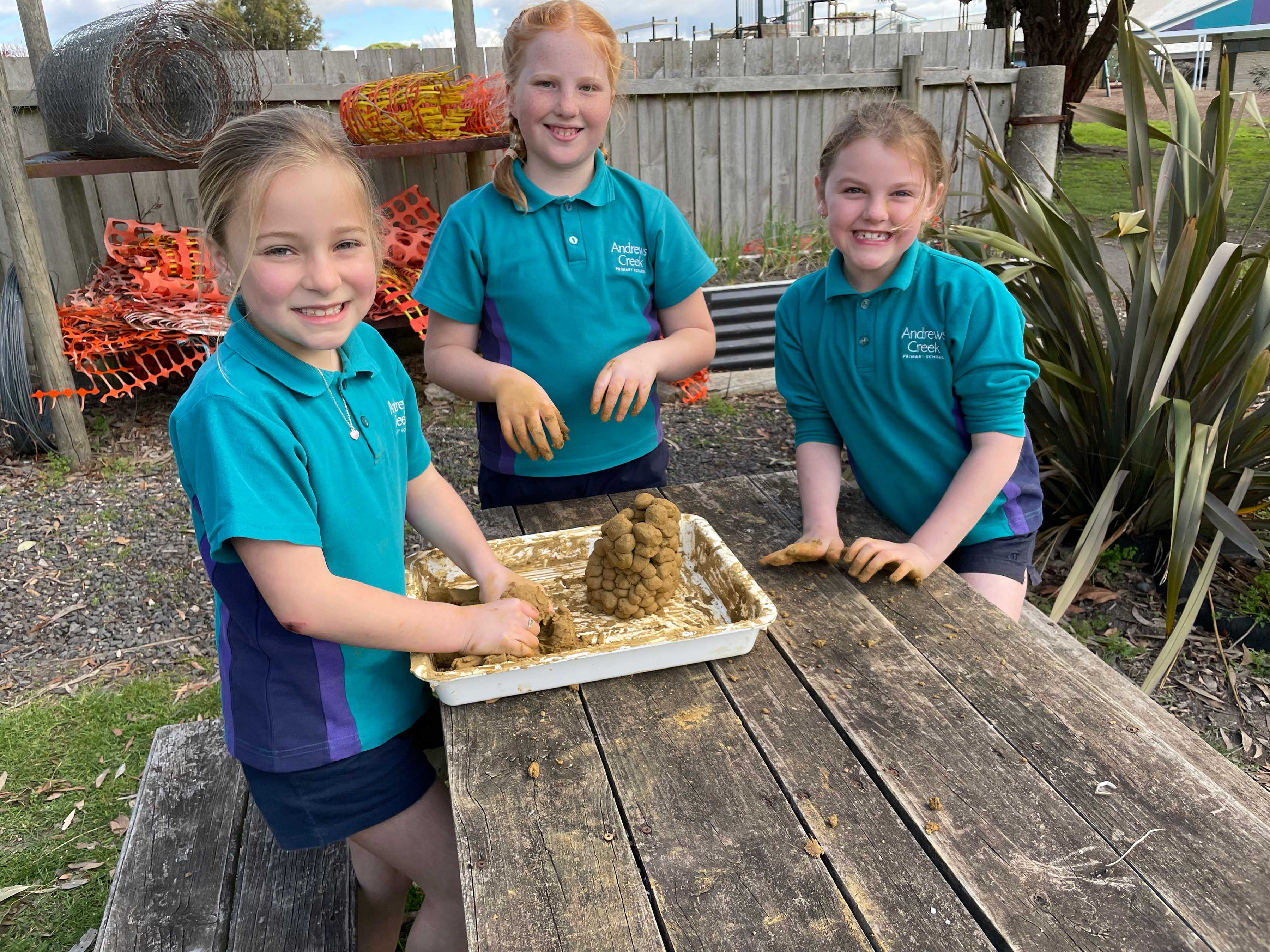 Three students from Andrews Creek Primary School pose with a model of a burrowing crayfish chimney which they have just created out of clay, in a white tray on a wooden picnic table, in an outside garden area. Photo: Clare Hawkins.