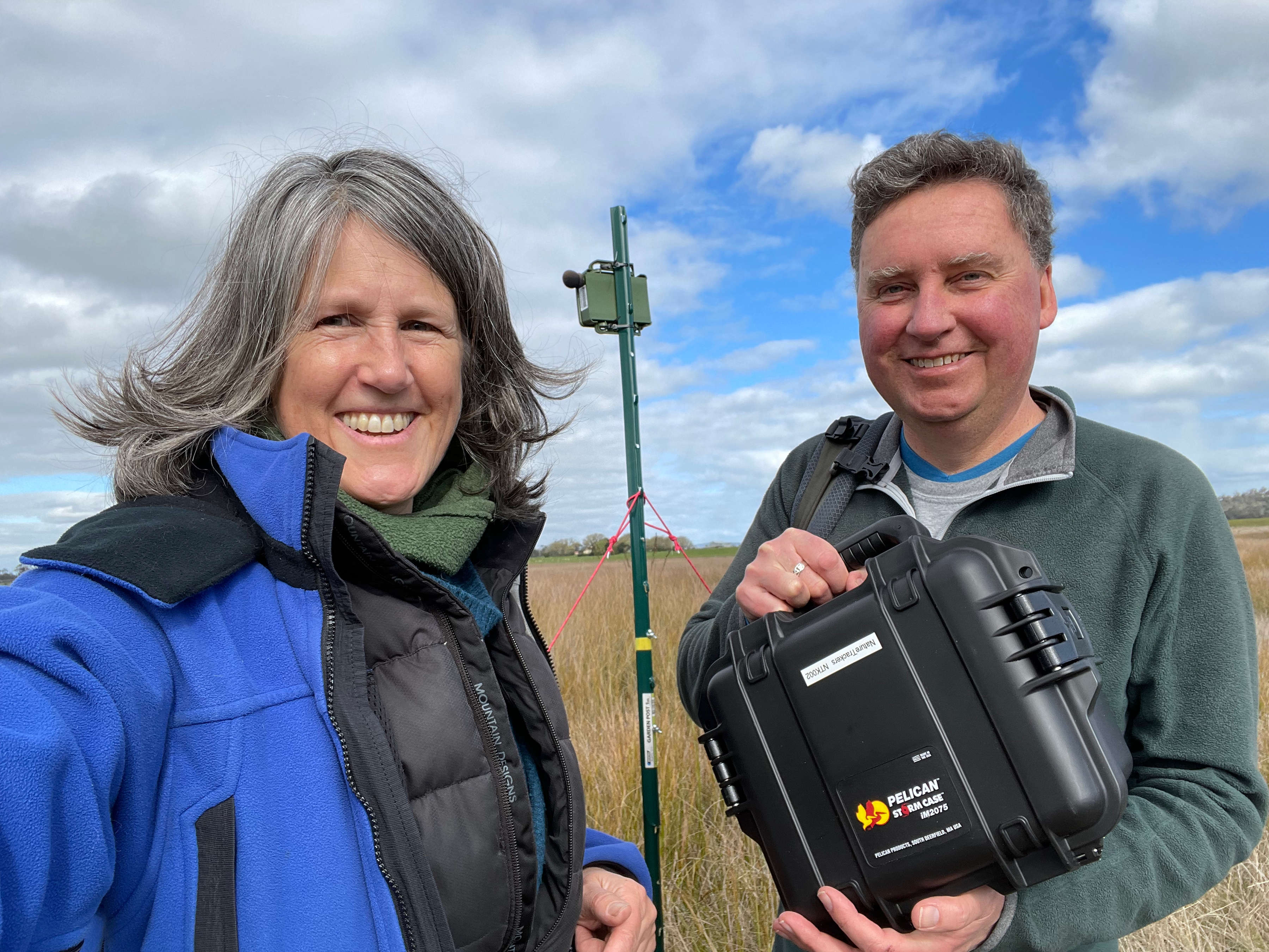 Clare Hawkins and Jim Lovell standing in a field of grass on a cloudy day. They are looking directly at the camera and smiling. Jim is holding a small black equipment case. The person on the right has their arm extended, taking the selfie. In the background between them is a pole with a small green box attached at the top. They are tracking wedge tailed eagles.