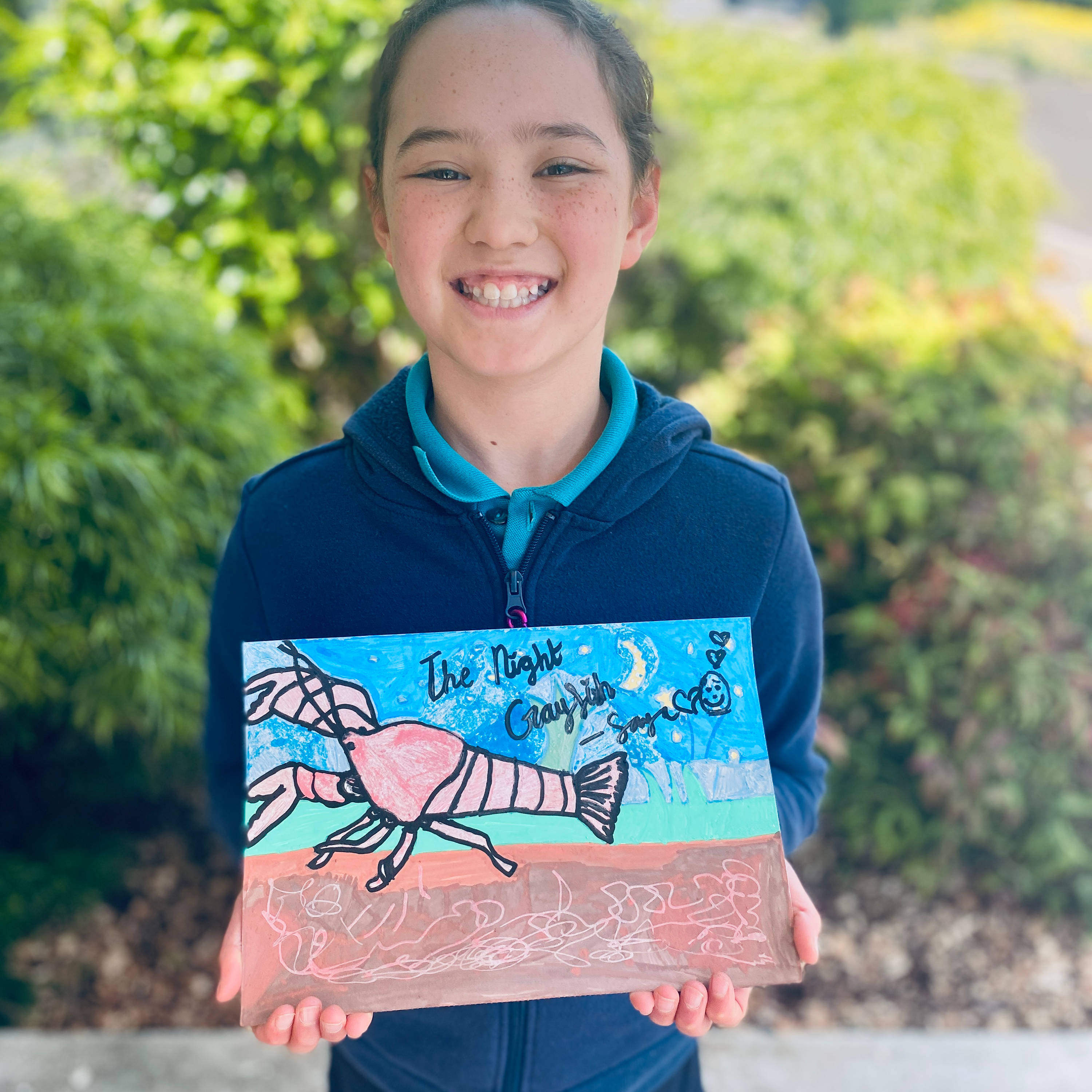 A primary school student wearing a blue zipped hoodie and a large smile. They are holding in front of their chest their drawing of a Tasmanian burrowing Crayfish.