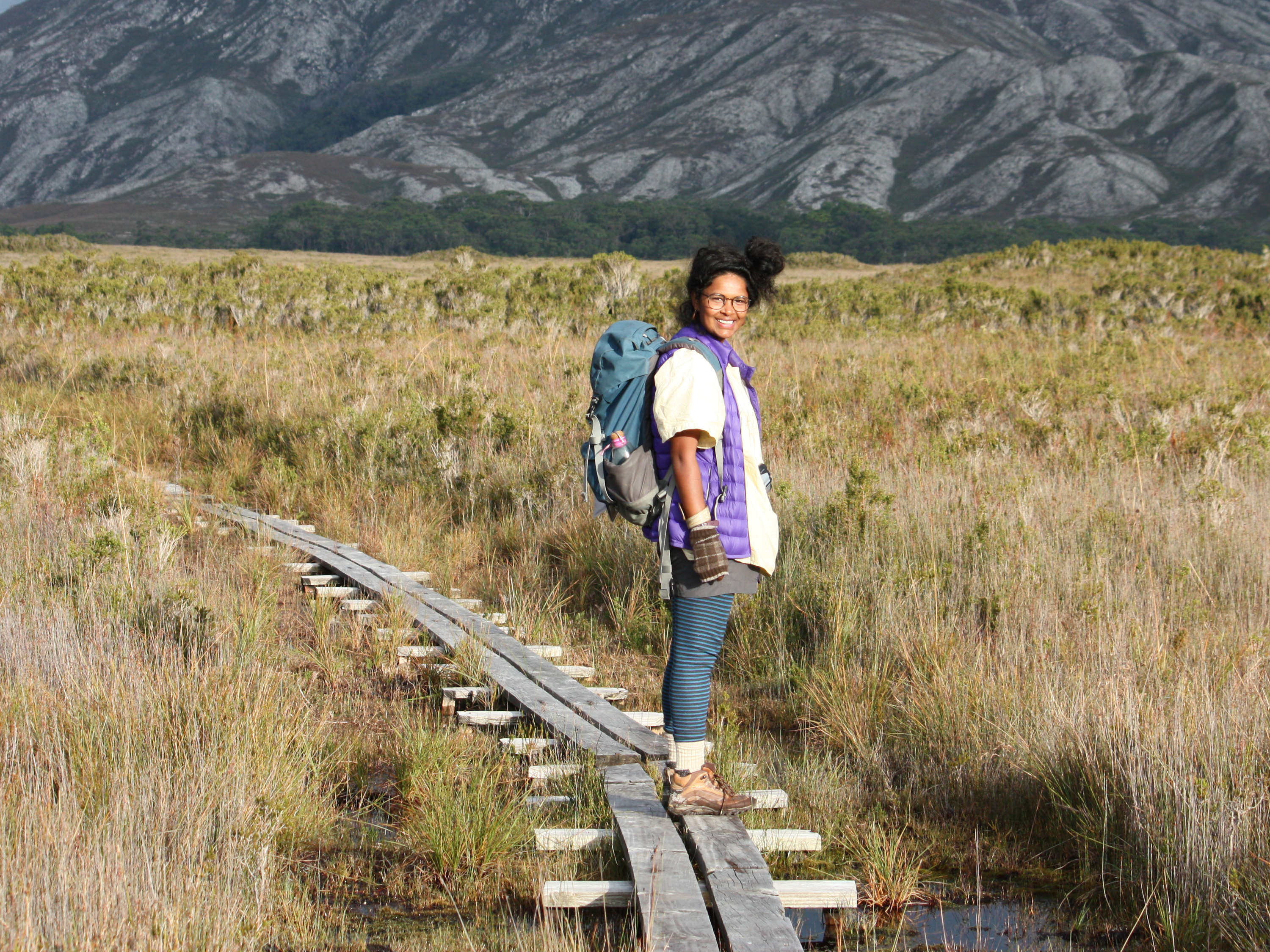 A woman stands on a boardwalk in the sunshine, in grassland in Tasmania’s remote southwest, with a mountain behind her. She is smiling at the camera. Photo: Persia Brooks.